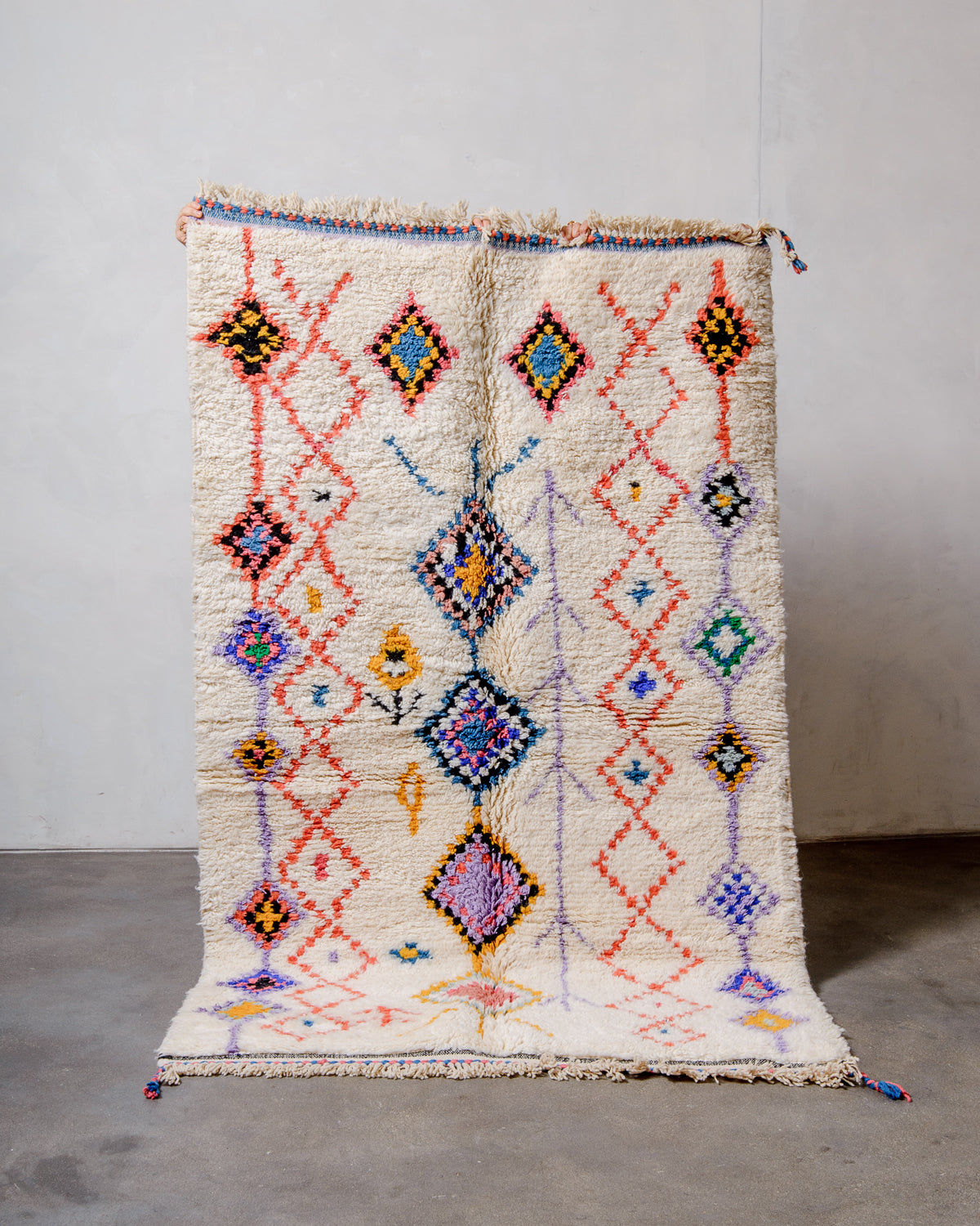 Berberlin Beniourain rug in colourful wool and cream, being held up in front of a white wall.